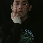 A young man standing in front of a solid black background. Rainbow tears are graphically drawn coming out of the right eye, and there are blue tears coming out on the left. Model is posed in a neutral stance with middle finger pulling down the eye with blue tears. "F*g" is written on his face.