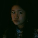 An image of a woman with blue gemstones under and over her eyes. She is standing in front of a pitch black background. She is looking straight towards the camera and the light.