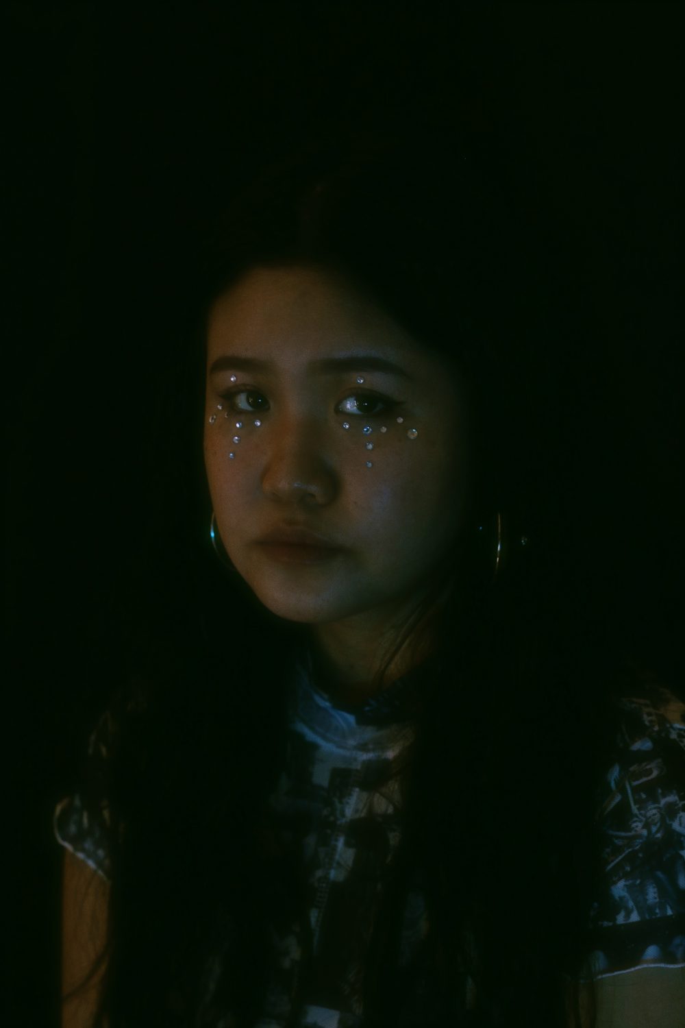 An image of a woman with blue gemstones under and over her eyes. She is standing in front of a pitch black background. She is looking straight towards the camera and the light.