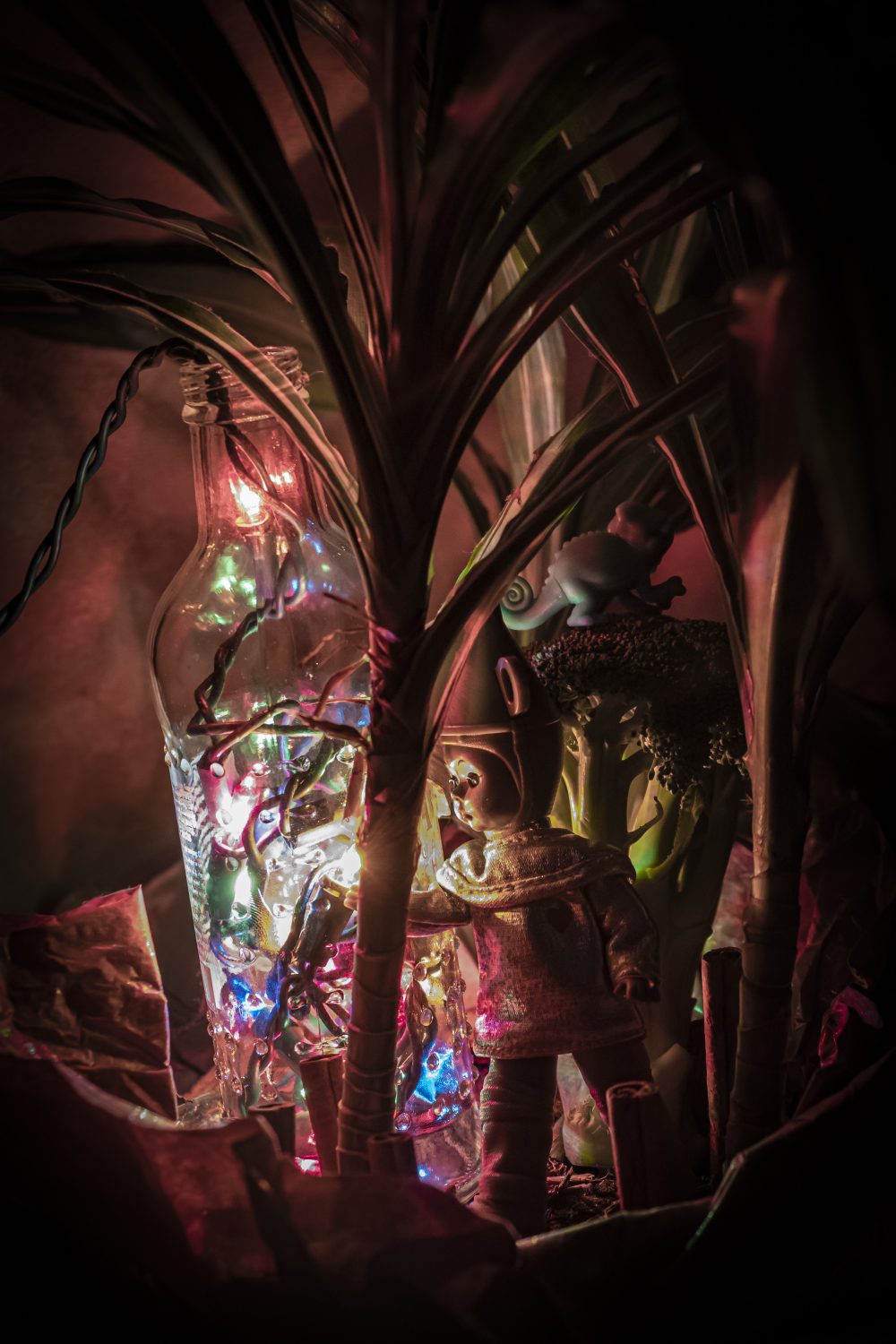 A constructed photo displaying a doll inside of a potted plant with all sorts of random objects like broccoli, cinnamon sticks, another objects that might not be seen in frame. The lighting of the image is used from rainbow Christmas lights in a glass bottle.
