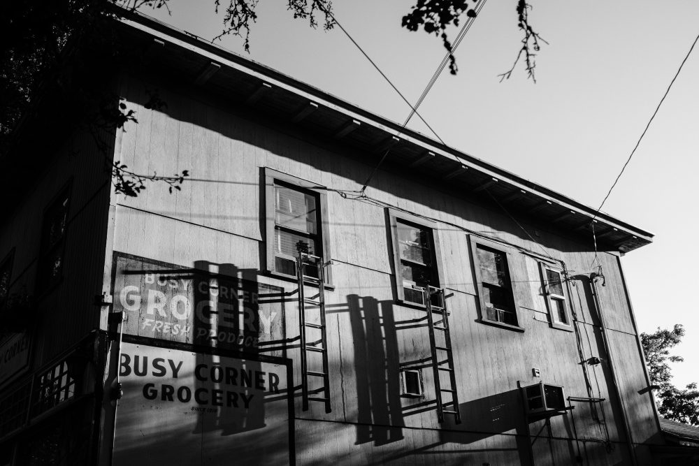 This is a black and white image of an exterior wall of an old grocery building. The perspective is from below, looking up at the old, weathered wall with signage that reads "Bust Corner Grocery". There are old iron fire escape ladders hanging from two windows and casting a shadow on the building.