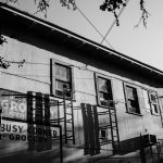This is a black and white image of an exterior wall of an old grocery building. The perspective is from below, looking up at the old, weathered wall with signage that reads "Bust Corner Grocery". There are old iron fire escape ladders hanging from two windows and casting a shadow on the building.