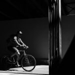 This is a black and white photo of a man on a bicycle under a bridge. He is illuminated by a beam of light that shines through a gap in the bridge as he rides along the path.