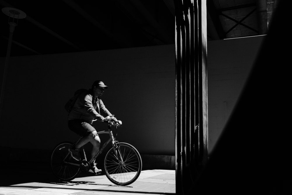 This is a black and white photo of a man on a bicycle under a bridge. He is illuminated by a beam of light that shines through a gap in the bridge as he rides along the path.