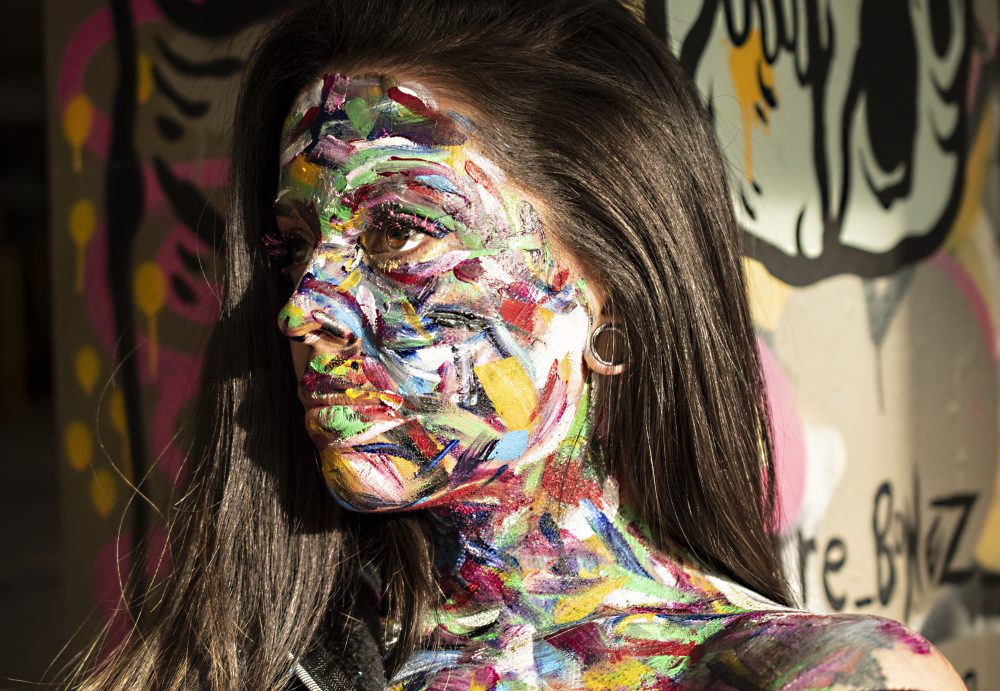 Model with bright paint all over her face and neck taken in front of graffiti.
