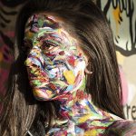 Model with bright paint all over her face and neck taken in front of graffiti.