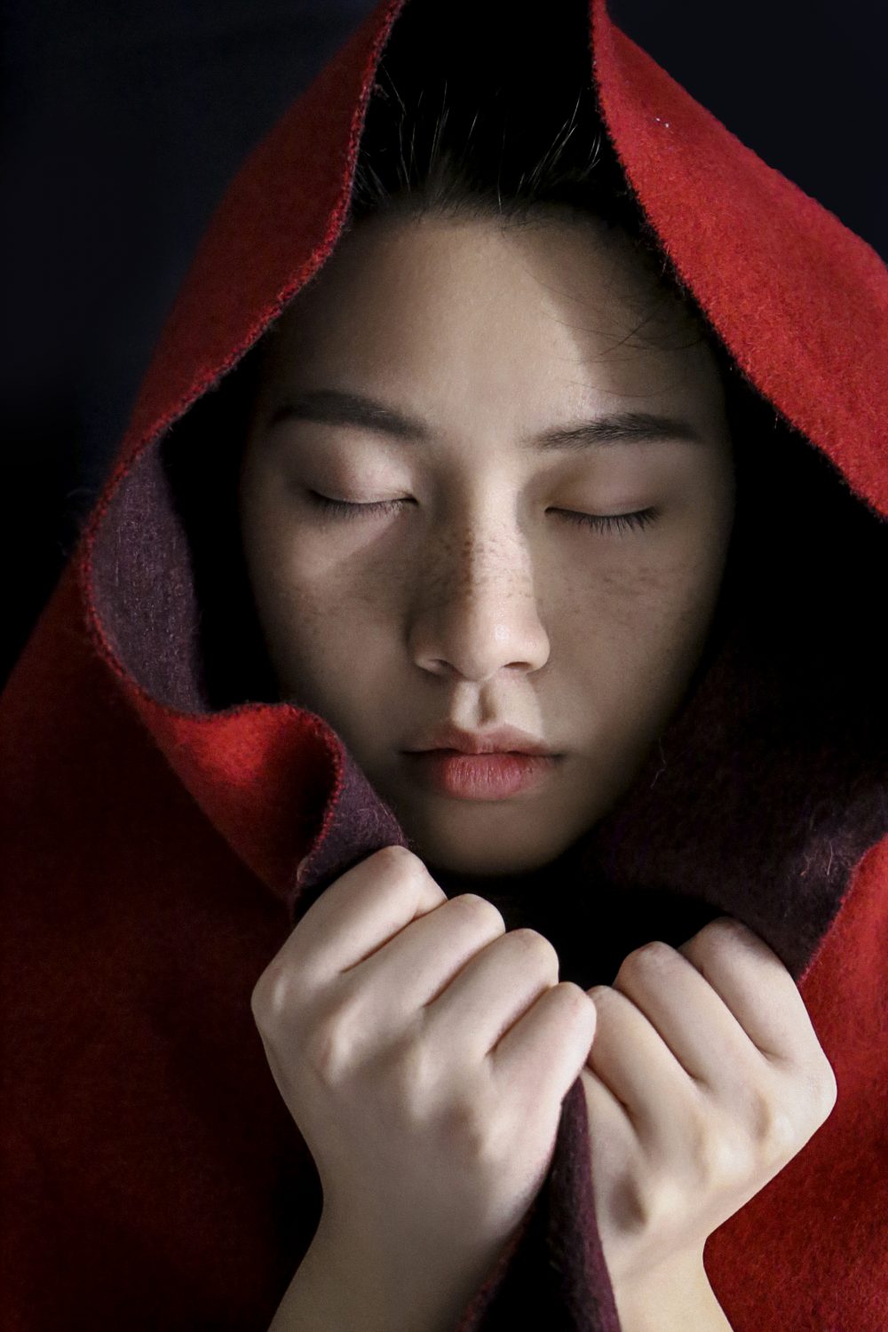 Woman wearing a read blanket over her head with her eyes closed and hands holding the blanket.