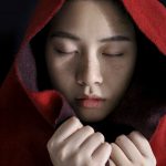 Woman wearing a read blanket over her head with her eyes closed and hands holding the blanket.