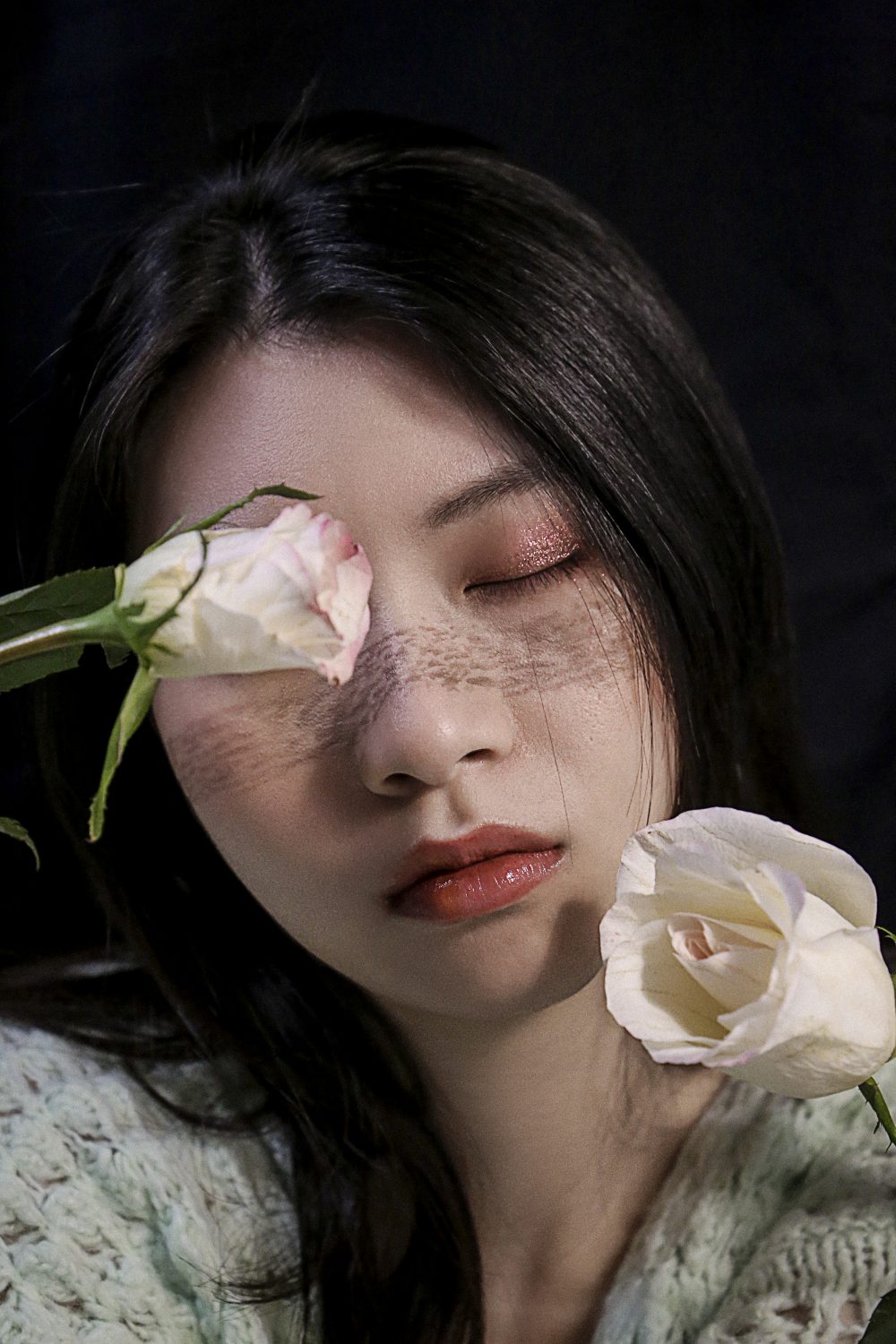 Woman in front of a black background, holding two white and pink roses with one held in front of her right eye.