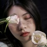 Woman in front of a black background, holding two white and pink roses with one held in front of her right eye.