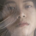 Photograph of a woman looking at the camera with a thin layer of white cloth in front of her face.