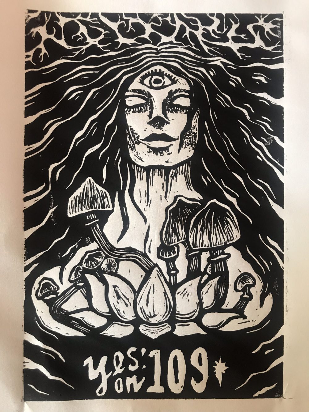This is a black and white relief print. There is a woman's face with her eyes closed and a third eye on her forehead that is open. Above her head is a representation of firing synapses, which appear like lightening. Her hair is long and black with white highlights, and it becomes the background of the image. Below her face are Psilocybin mushrooms growing up from behind a lotus flower. Under the flower, text reads "Yes! on 109".