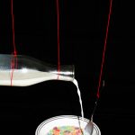 A bowl of cereal and a glass of milk held up by bright red strings, with the milk pouring in to the bowl. The images are surrounded by black so they appear to float in space.