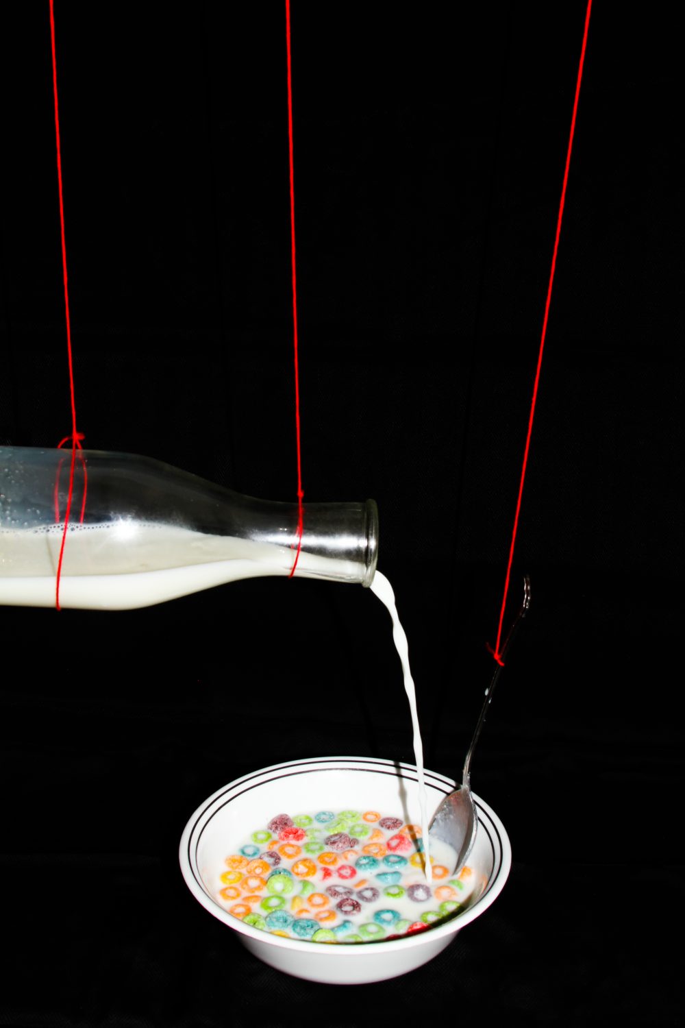 A bowl of cereal and a glass of milk held up by bright red strings, with the milk pouring in to the bowl. The images are surrounded by black so they appear to float in space.