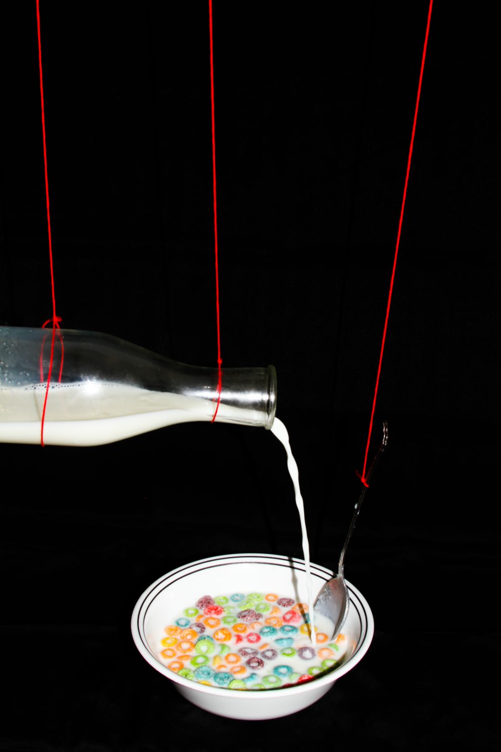 A bowl of cereal and a glass of milk held up by bright red strings, with the milk pouring in to the bowl. The images are surrounded by black so they appear to float in space.