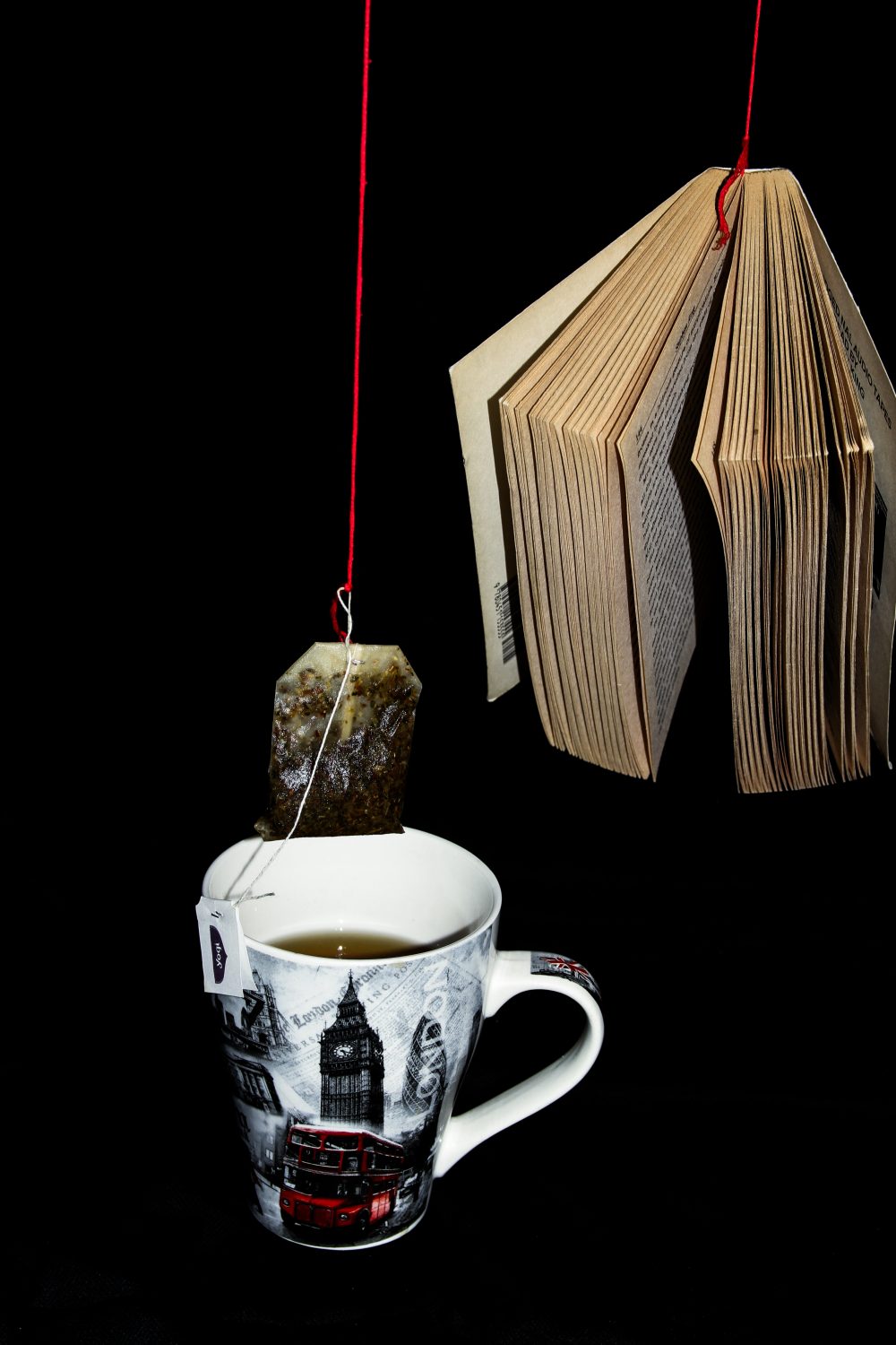 A book held up in empty space with bright red string, next to a cup of tea and teabag being held up by bright red string as well