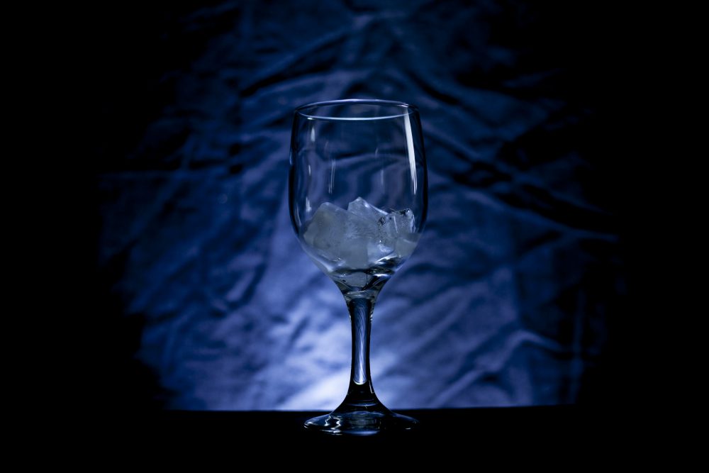 A wine glass with ice cubes in front of a drop-cloth that is lit up with blue light.