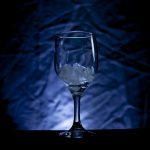 A wine glass with ice cubes in front of a drop-cloth that is lit up with blue light.