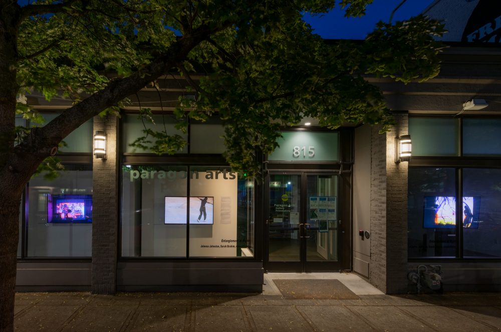 Window view of Paragon with three videos playing on monitors at night.