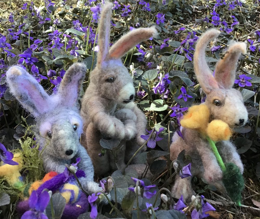 Three small felt bunnies in a field of violets holding flowers and a basket.