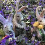 Three small felt bunnies in a field of violets holding flowers and a basket.