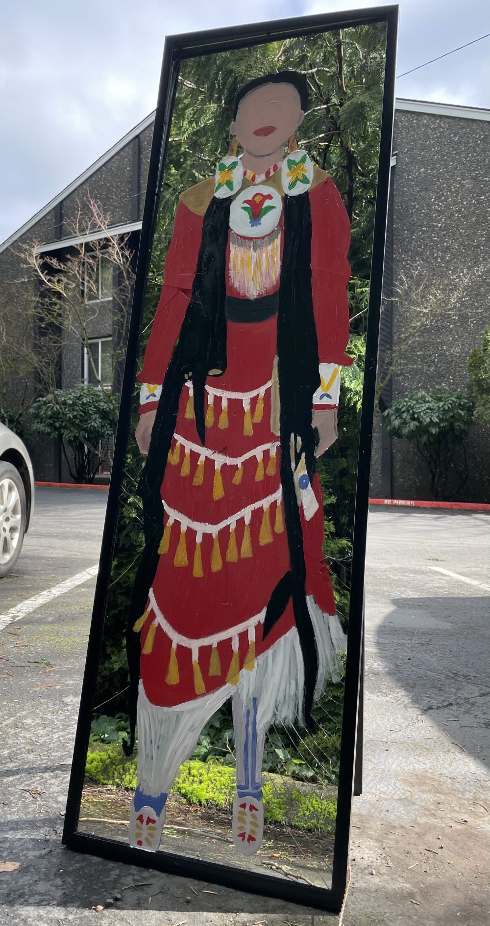 A painting of a jingle dress dancer on a shattered mirror. The woman's long dress is red, with many gold accessories. Around the woman, in the reflection of the mirror, nature is visible.