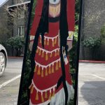 A painting of a jingle dress dancer on a shattered mirror. The woman's long dress is red, with many gold accessories. Around the woman, in the reflection of the mirror, nature is visible.