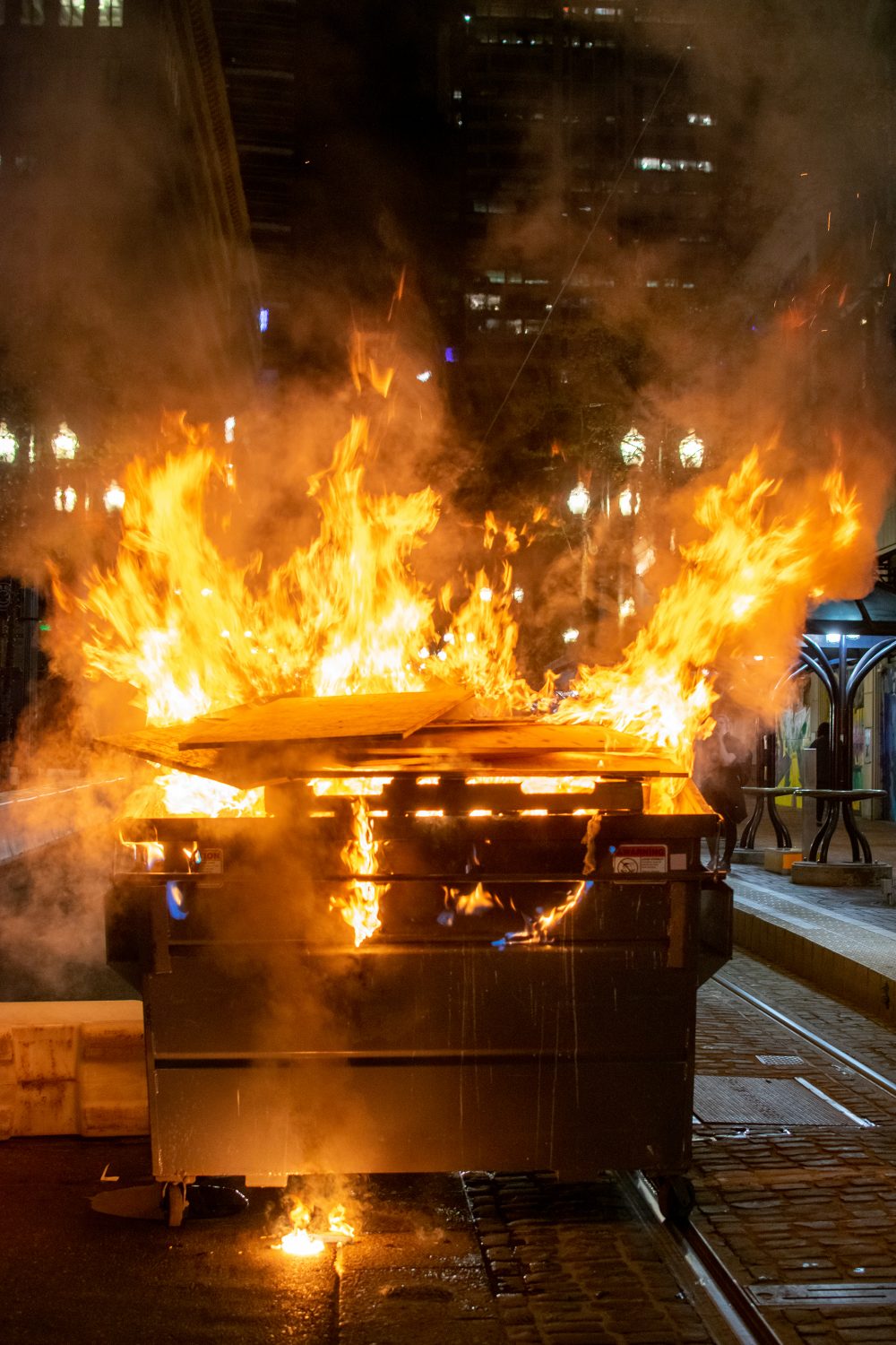 Image shows a garbage dumpster, which is on fire, blocking the middle of a street in downtown Portland.