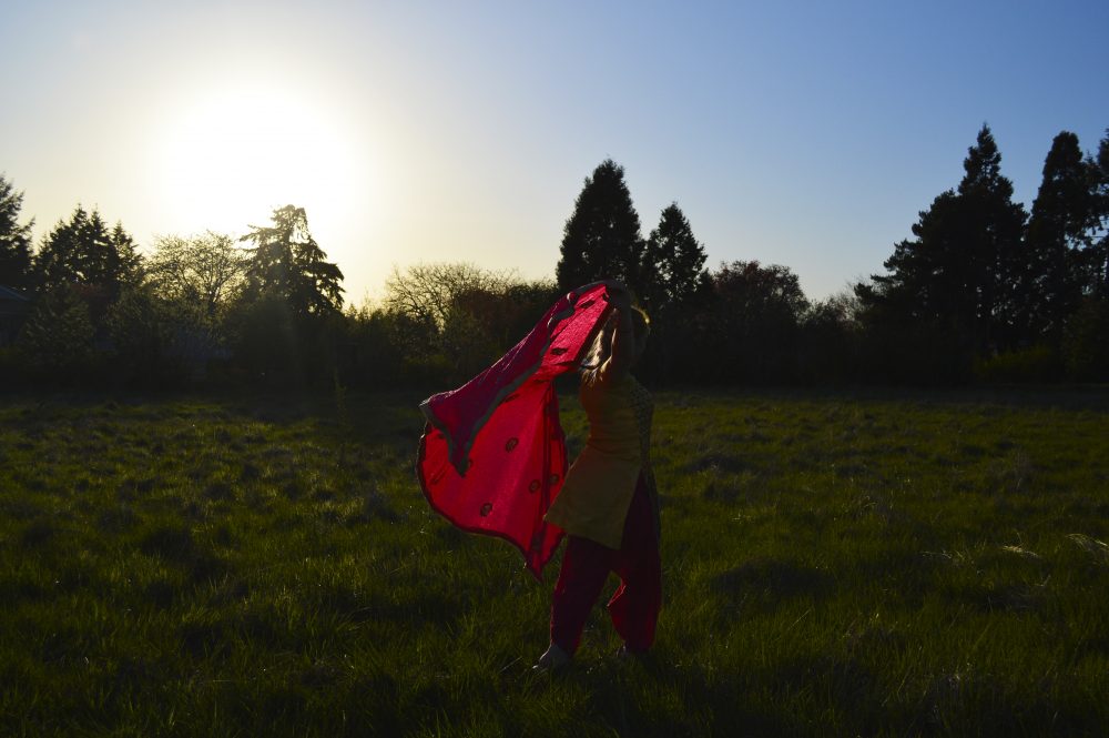 A human walking in a grassy field, holding a bright red fabric which is floating behind the human.
