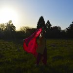 A human walking in a grassy field, holding a bright red fabric which is floating behind the human.