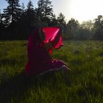 Human in grassy field holding large red fabric up to the sun.