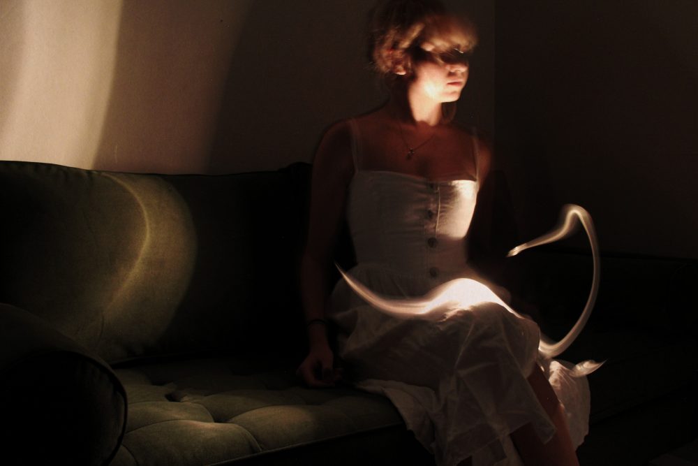 A photograph of a girl sitting on the left side of a moss green couch.There is a snake-like light source gliding across her lap in the foreground, and another light reflecting on the far right of the green couch and the white wall in the background.