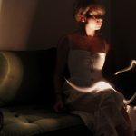 A photograph of a girl sitting on the left side of a moss green couch.There is a snake-like light source gliding across her lap in the foreground, and another light reflecting on the far right of the green couch and the white wall in the background.