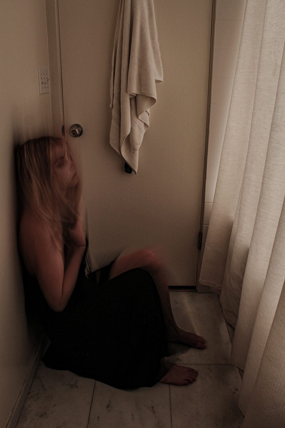 A photograph capturing the movement of a woman in distress standing in a bathroom near the door in a long, black prom dress, sliding her back down the white bathroom wall to sit on the white marble floor.