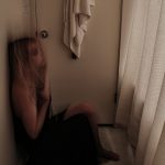 A photograph capturing the movement of a woman in distress standing in a bathroom near the door in a long, black prom dress, sliding her back down the white bathroom wall to sit on the white marble floor.