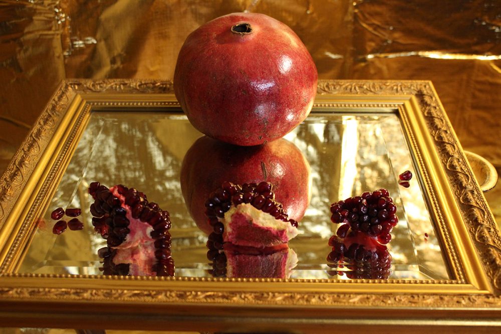 A photograph of a whole pomegranate and three pomegranate pieces served onto a mirror with a gold background.