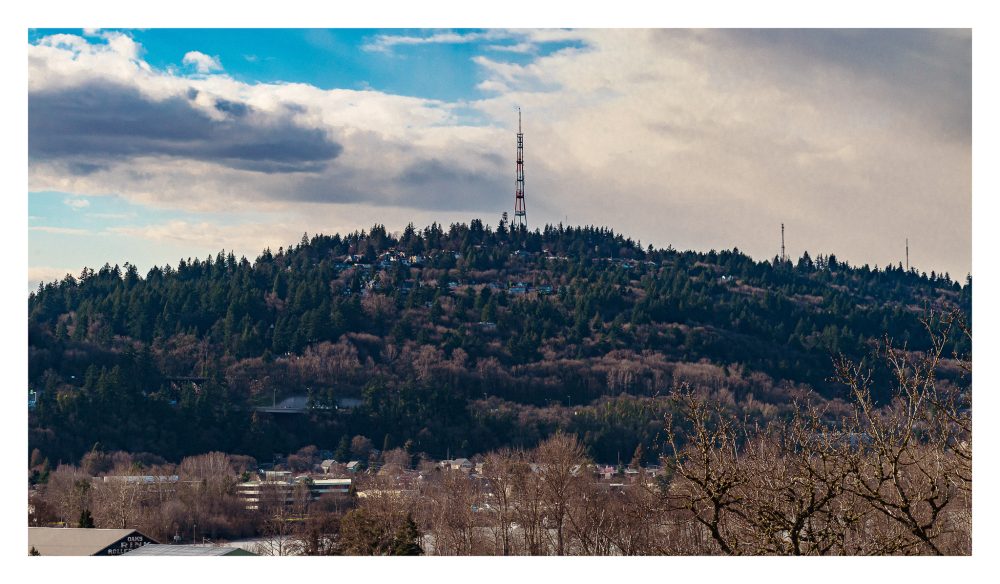 A landscape photograph of rolling hills covered in trees and houses, topped by three stoic radio towers repeated into the distance.