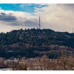 A landscape photograph of rolling hills covered in trees and houses, topped by three stoic radio towers repeated into the distance.