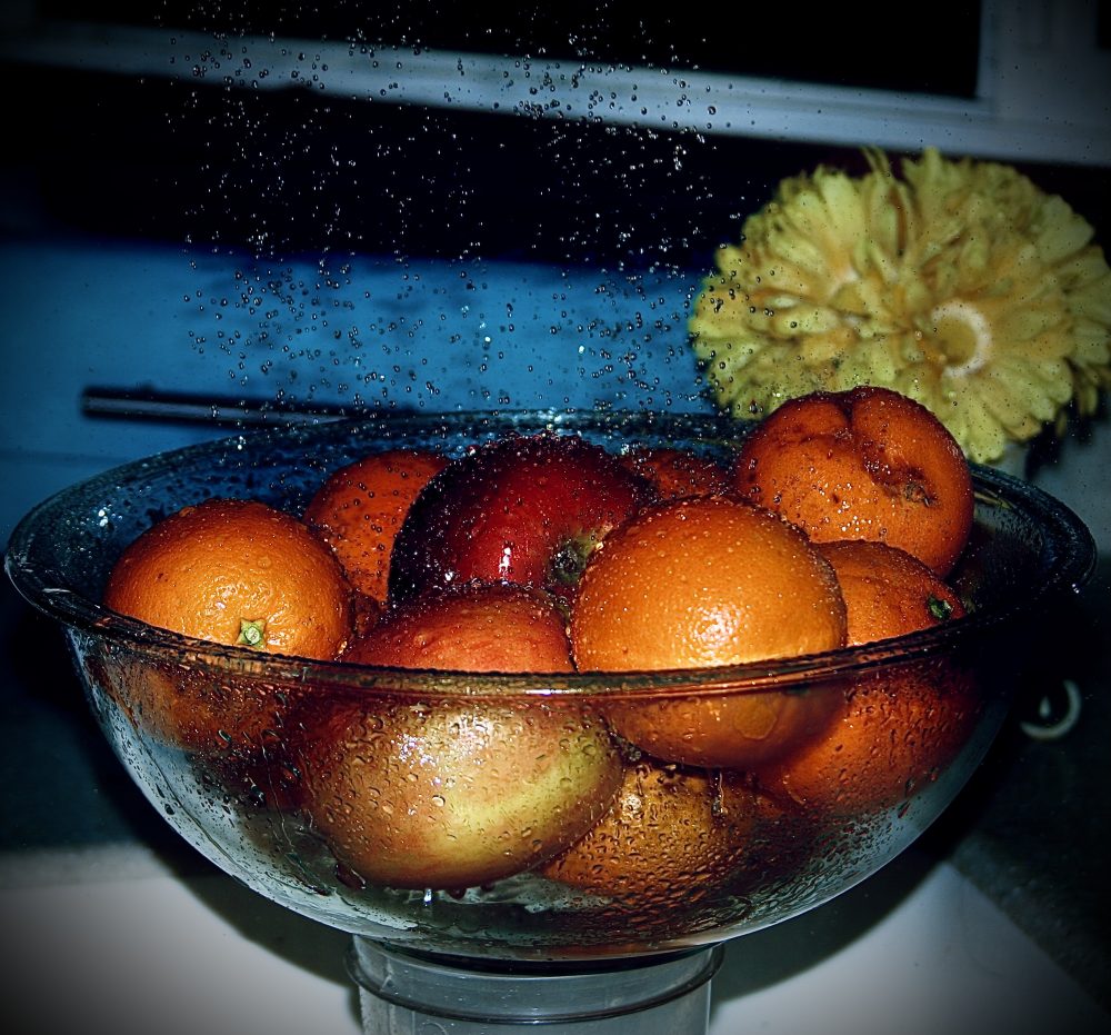 A photograph of water droplets being showered onto fruit for a nice cleaning. With a flower pot and a blue bread basket to show more vibrant colors.