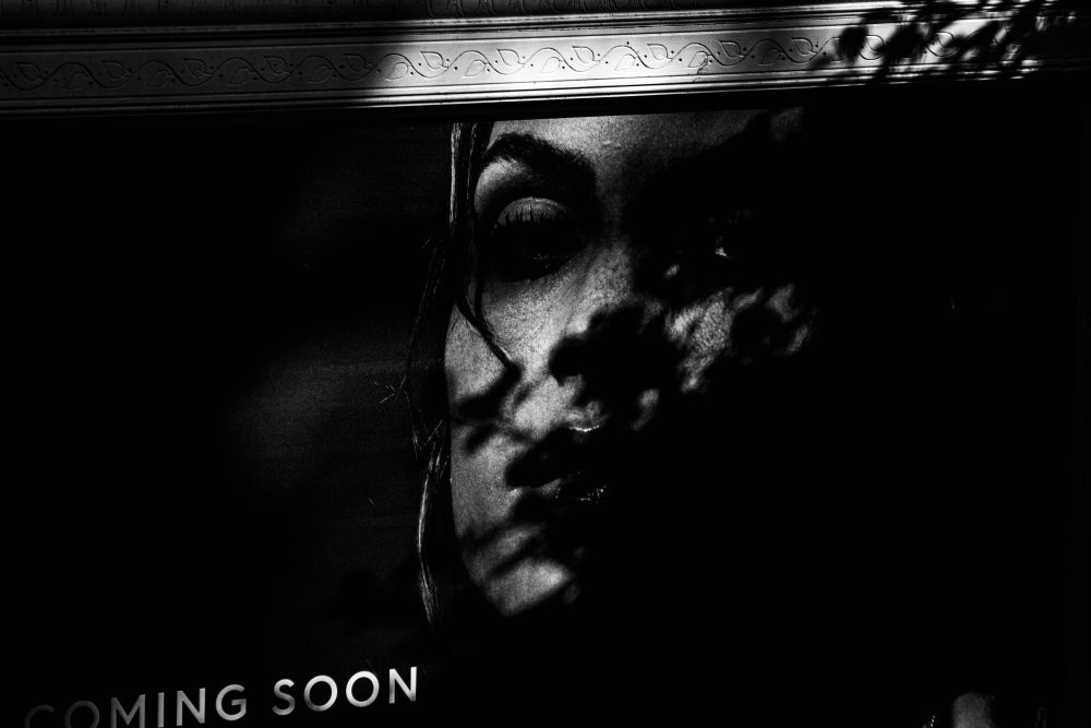 Black and white photograph of a commercial poster of a woman's face, partly covered by the shadow of a tree branch.