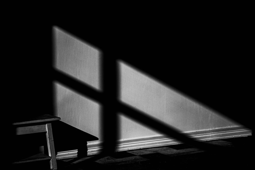 Photograph of a shadow cast from light shining through a window cast upon a low sitting stool stretched out onto a checkered floor.