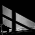 Photograph of a shadow cast from light shining through a window cast upon a low sitting stool stretched out onto a checkered floor.