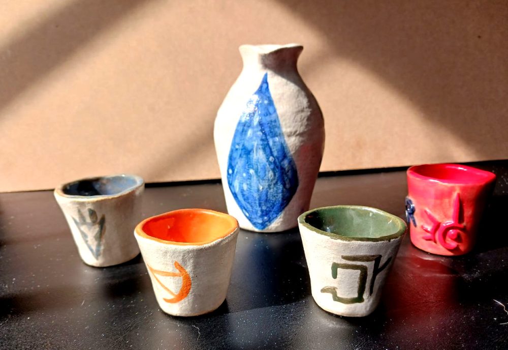 A colorful sake set, white sake flask with a blue crystal painted on it, four small cups to accompany it with various FFXIV class symbols.