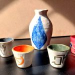 A colorful sake set, white sake flask with a blue crystal painted on it, four small cups to accompany it with various FFXIV class symbols.