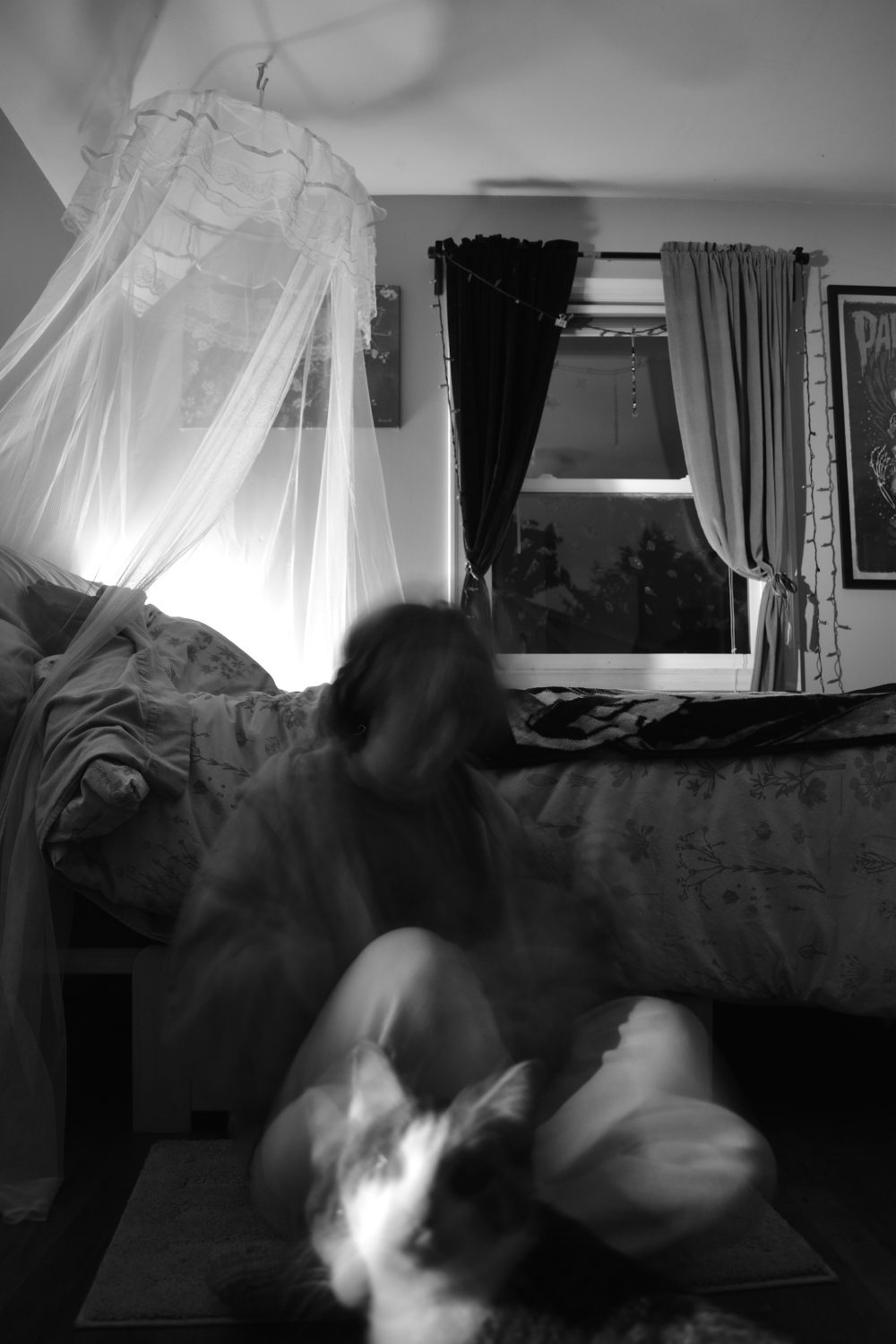 A black and white image of a moving figure sitting in front of a messy bed. A blurry cat is walking in front of the figure.
