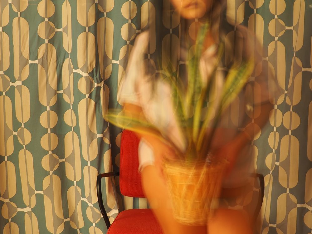 A ghostly woman figure stands in front of a red chair holding a plant. She has long black hair and a neutral facial expression. The background is of blue and white pattern curtain.