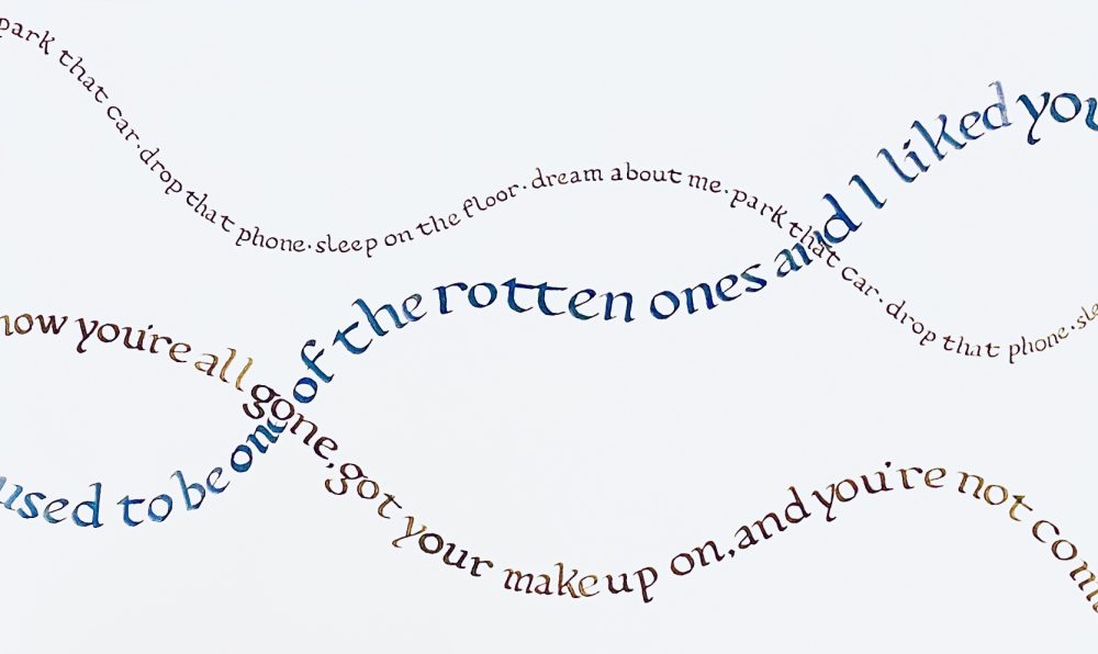 This is a piece of paper with song lyrics written in calligraphy in three sizes, all intersecting and written on curves, like hills, in blue gray and ochre colored inks.
