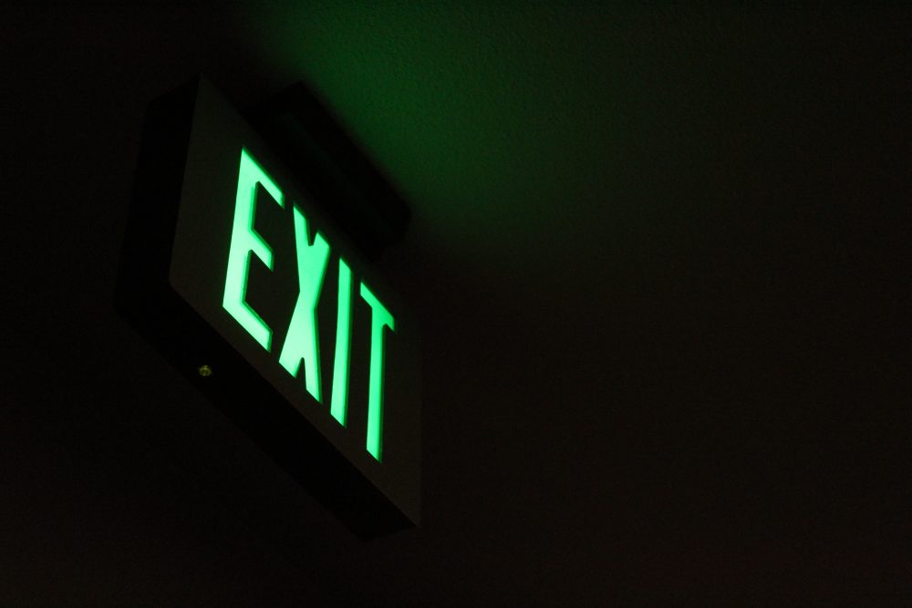 The pale green light of an exit sign stands against a mostly black background. A faint glow from the sign defines a vague sense of a room.