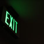 The pale green light of an exit sign stands against a mostly black background. A faint glow from the sign defines a vague sense of a room.