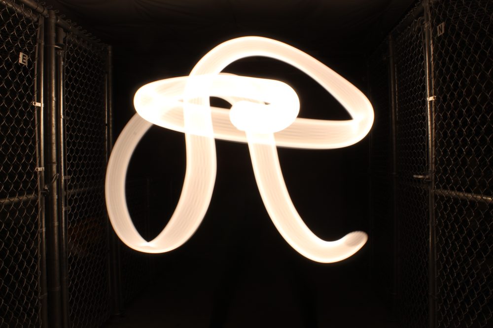 In a dark room, a streak of light forms a swirling script letter R to either side a chain link fence is lightly illuminated by the glow.